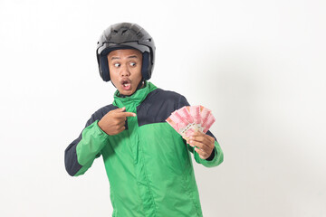 Portrait of Asian online taxi driver wearing green standing against white background, holding a bunch of rupiah money while pointing to the side
