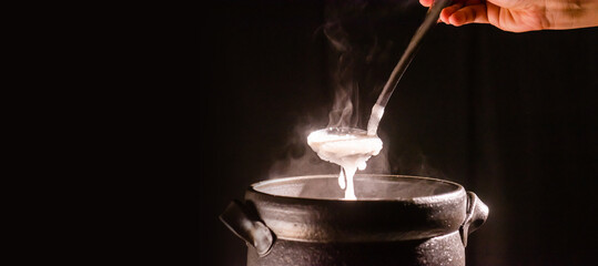 The cook was cooking rice porridge in a boiling clay pot with steam coming out and using a ladle to scoop the rice porridge.