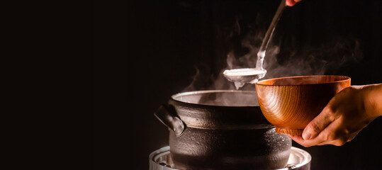 The cook was cooking rice porridge in a boiling clay pot with steam coming out and using a ladle to...