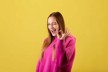 Portrait of a winking young woman making OK sign with fingers and looking at the camera over yellow...