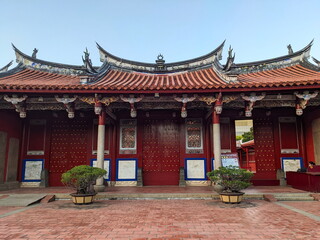 Fototapeta premium Tainan, Taiwan - 11.29.2022: Entrance to the main area of Tainan Confucius Temple with tiled roof and a ticket stall and a staff member on the right hand side under a blue sky during the pandemic