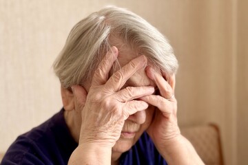 A close-up of the head of serious mature old woman sitting at home on the couch and holding her...