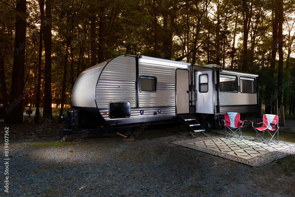 Wall mural Campsite with a large trailer and chairs in a forest with the sun low behind and colorful.