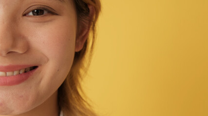 Close-up, half face of young woman looking at camera with smile isolated on yellow background in...