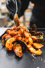 Close-up of a chef’s hands in black gloves serving spicy grilled shrimp curry on a plate, with a...