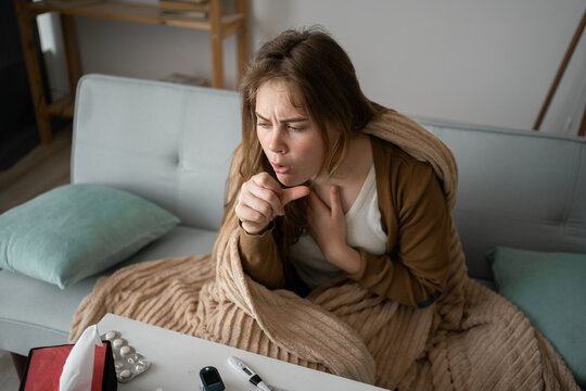 young sick upset woman sitting on the sofa at home covered with a blanket coughing intensely, sick girl with flu symptoms. copy space.