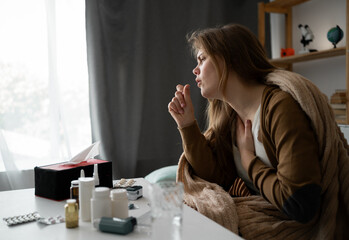 side view. young sick upset woman sitting on the sofa at home covered with a blanket coughing...