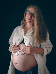 Portrait of a pregnant woman with glasses holding small baby socks in her hands. Black background....