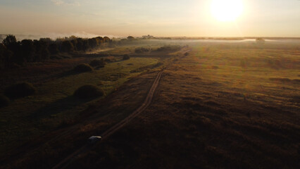 The car is driving along a country road at dawn