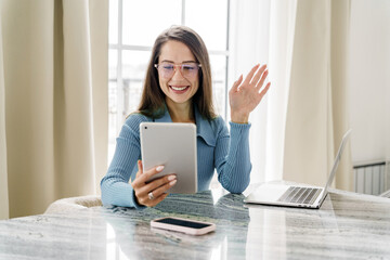 A professional woman with glasses, wearing a blue sweater, waving during a video call on a tablet.