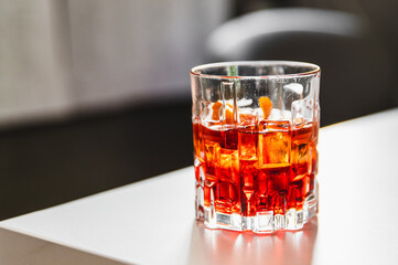 Amber-colored cocktail in a clear glass with ice cubes, placed on a surface, against a blurred background.