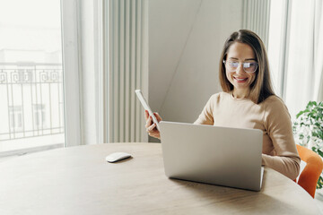 A professional woman with glasses, wearing a beige sweater, multitasking with a laptop and tablet.