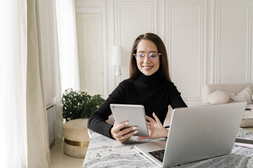 A professional woman with glasses, wearing a black sweater, using a tablet while working at a laptop.