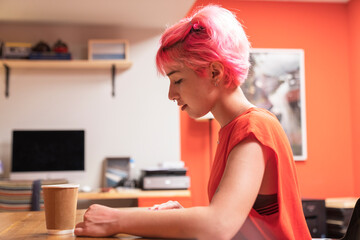 young woman working while drinking coffee in the office. Generation Z woman with pink hair works in...