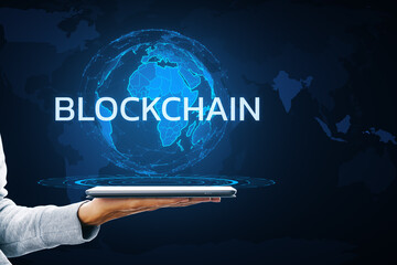 A person's hand holding a tablet displaying a holographic globe with the word BLOCKCHAIN on a dark background with map outlines, illustrating a digital concept