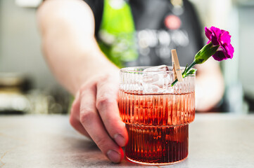 Close-up of a hand holding a glass of a reddish beverage, garnished with a purple flower, on a bar counter.