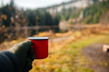 A guy holds a red mug on the riverbank in autumn