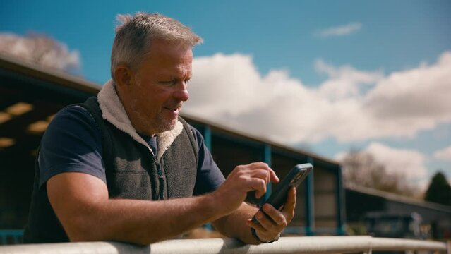 Mature male farm worker standing outside barn leaning on fence making call on mobile phone - shot in slow motion