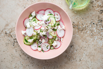 Roseate bowl with cucumber, radish and feta cheese salad, top view on a beige granite background,...