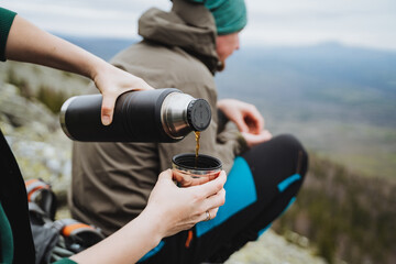 A traveler wearing a hat is pouring water from a thermos into a cup while enjoying the scenic...
