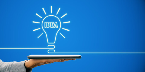 Neon light bulb with 'IDEA' text above a tablet held by a person, against a blue background,...