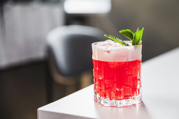 Refreshing red cocktail with ice and mint garnish in a glass, placed on a white surface, possibly a hibiscus mojito.