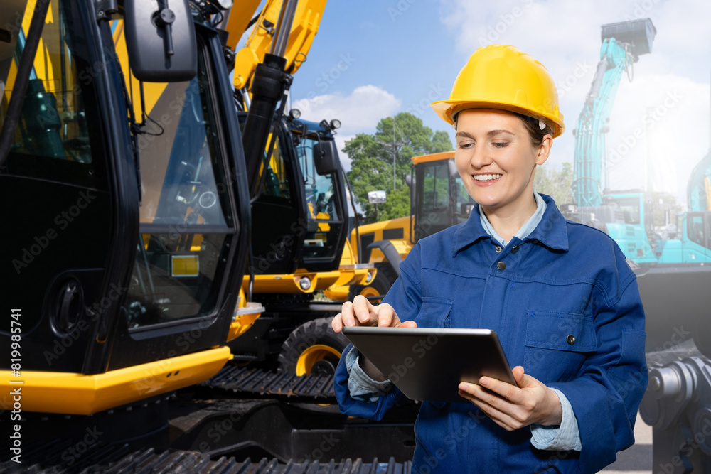 Poster female engineer in a helmet with a digital tablet stands next to construction excavators..