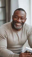 Smiling man with gray beard wearing a beige long-sleeve shirt holding a book sitting in a room with a blurred background.