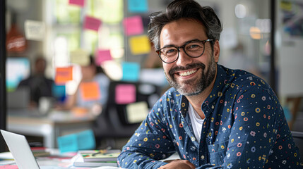a cheerful professional man working at his desk, with a laptop open in front of him and papers scattered around, a bright office environment with sticky notes on the glass partition in the background.