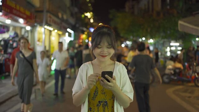 Portrait of happy Asian Vietnamese woman travel at night market, using mobile phone in technology device in people walking street fair in Hanoi city, Vietnam. Retail shops