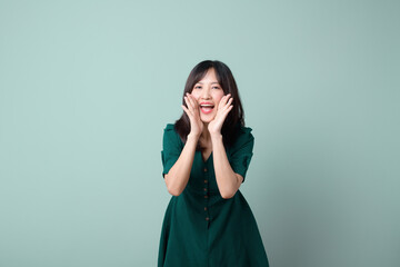 Happy Asian woman shouting above green dress poses against green background blank copy space for...