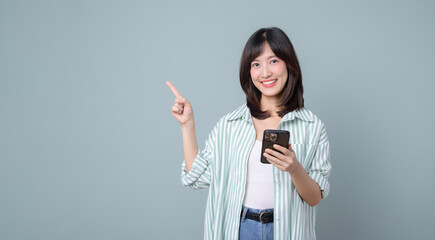 Excited asian girl shopping on mobile app, holding phone and pointing finger left at empty copy space, showing promo text, standing with smartphone against green background