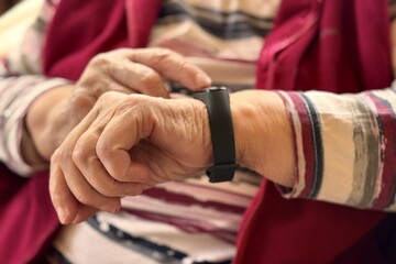 The black smart band with display touchpad and heart rate monitor. The old woman sitting in armchair touches and looks at the wristband of pulse monitor indoors. Fitness tracker