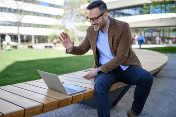 Successful manager waving hand while attending online meeting over laptop on bench in modern city