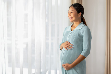 radiant Asian pregnant woman stands near a window draped with sheer curtains, gently cradling her...