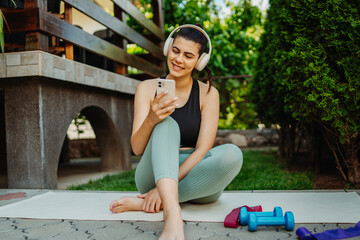 One young caucasian woman is sitting on yoga mat with wireless headphones and mobile phone preparing for training	
