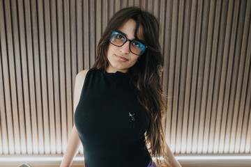 A stylish young woman wearing glasses stands confidently, sporting a chic black sleeveless top,...