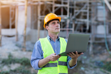Male caucasian technician engineer construction worker using computer laptop for digital blueprint architecture innovation analyzing planning building, wearing safety vest and helmet gears.