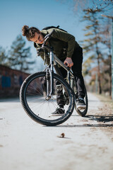A young man bends over his bicycle to fix it, equipped with a backpack in a serene park setting,...