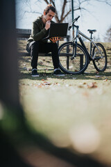A good looking young man engages with his laptop while sitting on a park bench, his bicycle parked...
