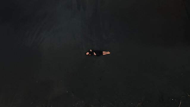 Aerial top down view of a beach with black sand on which a young attractive woman in a black dress lies, next to her are sea waves and foam. Depressed and sad mood on the beach, background video.