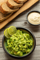 Chunky guacamole decorated with slices of lime, sourdough bread slices on an olive wood kitchen board, and tahini dip on the wooden table. Homemade, healthy vegan meal. 