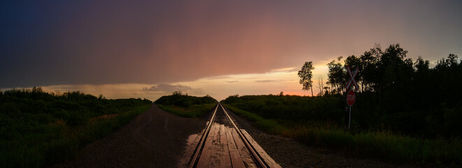 A late day view along a single set of railway tracks that disappear into the distance. The sky is mostly filled with gray to orange clouds. A recent rain has made the train tracks shine with the setti