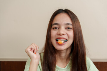 Young Asian girl with long brown hair eating orange slice, showing braces, smiling, and wearing...