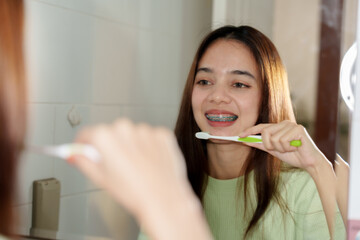 Asian teenage girl with long brown hair, wearing light green sweater, brushing teeth in front of...