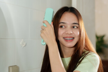 Young Asian girl with long brown hair, wearing light green sweater, brushing hair with teal comb,...