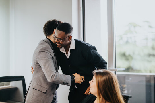 Two male business colleagues embrace in a well-lit office, showing support and camaraderie. A female coworker watches, contributing to the welcoming and inclusive work culture depicted.