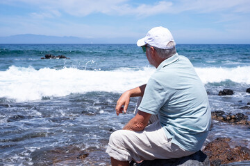 Back view of senior relaxed man barefoot wearing sunglasses and cap sitting on a rocky beach admiring the sea waves. Travel vacation leisure activity concept