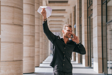 smiling man in a black shirt holds up papers while talking on the phone, walking through a modern office corridor with large pillars