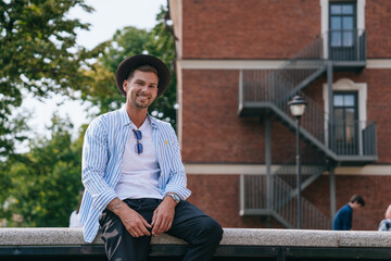 man in a striped shirt and hat sits on a ledge, smiling, in a park with a red brick building in the background - Powered by Adobe